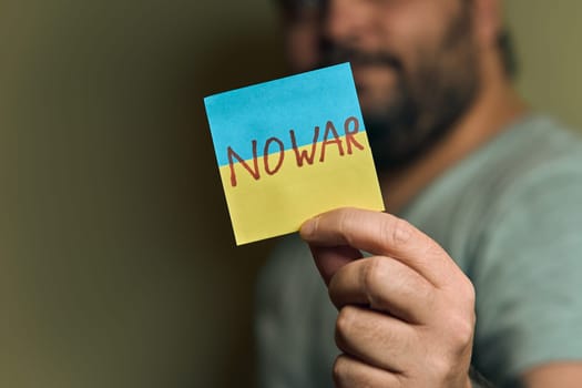 A bearded European man holds a sticker in front of him, in the form of a Ukrainian flag with the inscription no war