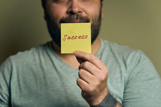A bearded European man holds a blue sticker in front of him, with the word success