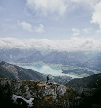 A vertical aerial shot of a young adult couple of hikers-photographers on top of the Austrian Schafberg mountain. Both in trekking equipment. The girl is standing sideways to the camera and facing the lake. The guy is sitting and controlling the drone. The grass on the mountain is of yellow autumn color. An alpine mountain range is in the background with clouds over it. Salzburg, Austria.