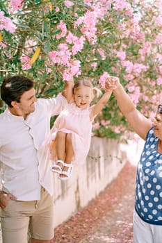 Dad and grandmother raise high by the arms a little laughing girl near a flowering tree. High quality photo