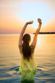 A young charming woman in a yellow dress stands on the seashore in front of a bright orange sunset. Romantic seascape