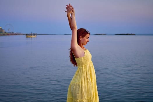Attractive young woman in a yellow dress posing with her arms raised above her head while waiting for the sunrise on the sea.