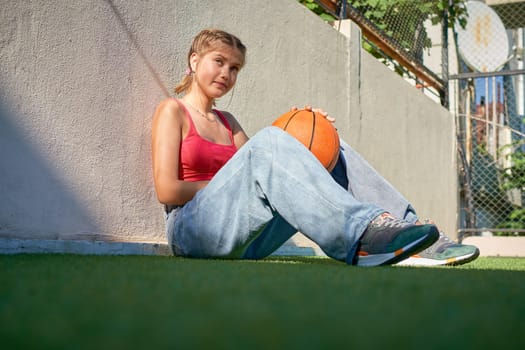 A girl sits with a basketball on the sports ground and laughs cheerfully