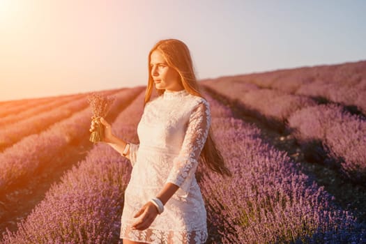 Close up portrait of young beautiful woman in a white dress and a hat is walking in the lavender field and smelling lavender bouquet.