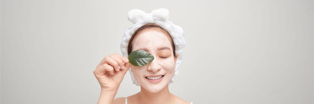 Smiling female hold fresh green leaf close to eye with  clay mask on her face.
