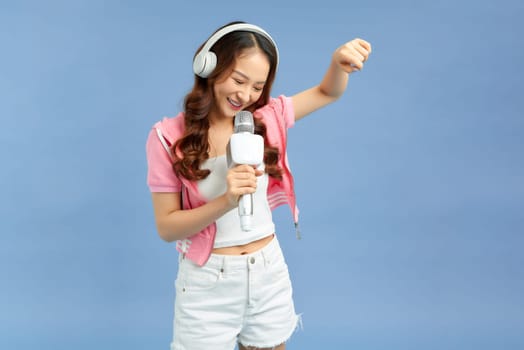 Optimistic young female singer in wireless headphones singing into microphone on blue background