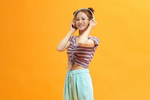 Beautiful woman in headphones listening music and smiling while standing against yellow background