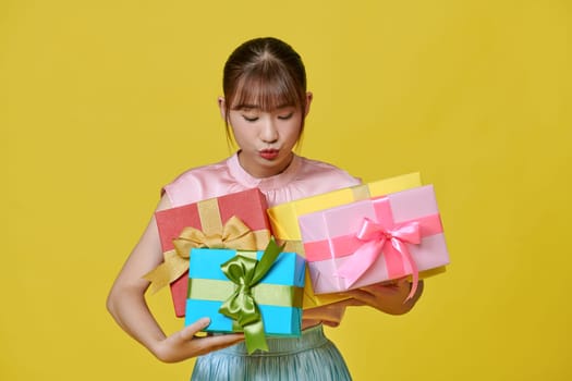 Attractive young girl in dress holding stack of gift boxes isolated over yellow background