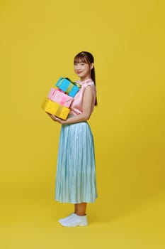 Attractive young girl in dress holding stack of gift boxes isolated over yellow background