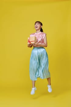 Happy pretty young woman jumping and holding wrapped gift box smiling isolated on yellow background