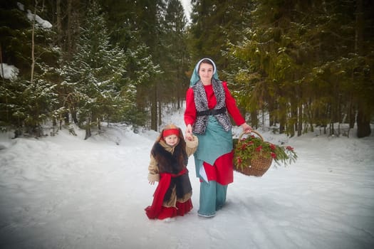 Family with mother and little daughter in stylized medieval peasant clothing in winter forest. The woman and child pose for fairy tale photoshoot in nature on a cold day. Concept of love, friendship