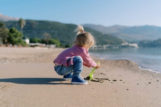 Little girl digs sand on the beach with a toy shovel. Side view. High quality photo