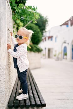 Little girl in sunglasses stands on a bench in the yard and looks at the wall. High quality photo