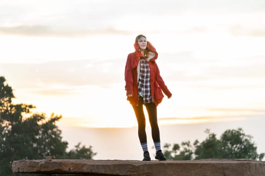 Beautiful Caucasian woman with red coat stand on the rock near the cliff on the mountain and background with warm light of sun rise in the morning.