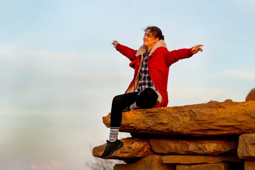 Beautiful Caucasian woman sit on the rock near cliff on the mountain and spread her arms with happiness and look forward with warm light in the morning.