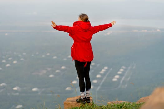 Close up back of woman wear red coat and stand with spread her arms near cliff on the mountain and look forward to beautiful view with warm light of sun rising.