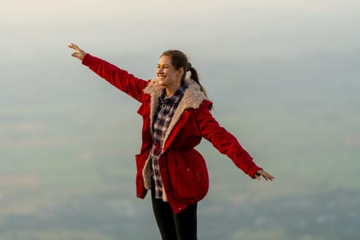 Caucasian woman stand near cliff with background of grass field and rural village area in view on the mountain with warm light of sun rise and she also look to left side.