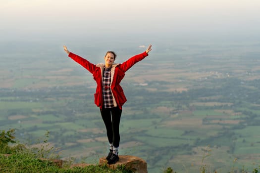 Wide shot of woman wear red coat and stand with spread her arms near cliff on the mountain and look at camera with beautiful high view background with warm light of sun rising.