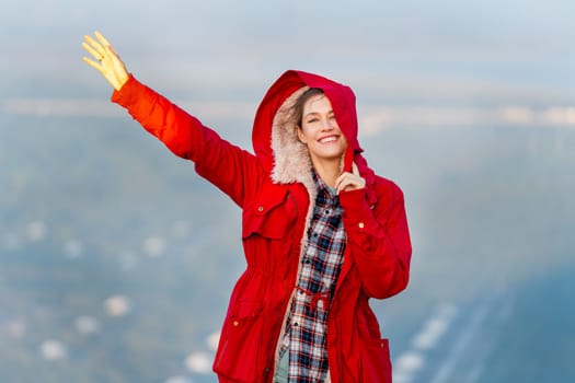 Beautiful woman stand near cliff with hold coat hood and wave hand on the mountain with warm light of sun rise and she look happiness with smiling also look at camera.