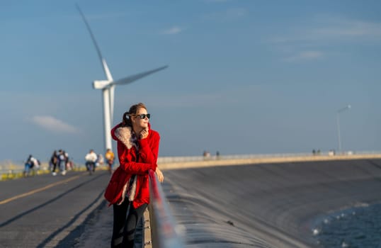 Beautiful Caucasian waman with sunglasses stand in front of wind turbine or windmill also look to right side near the road with smiling of happiness.