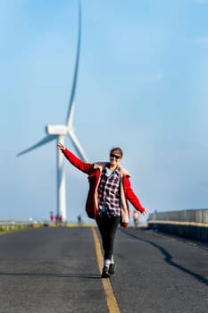 Vertical image of Caucasian woman enjoy to walk on single solid yellow line of the road on the mountain with big windmill or wind turbine on the back.