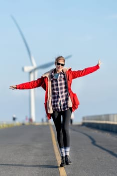 Caucasian woman enjoy to walk on single solid yellow line of the road on the mountain with big windmill or wind turbine on the back and she also smile to camera.