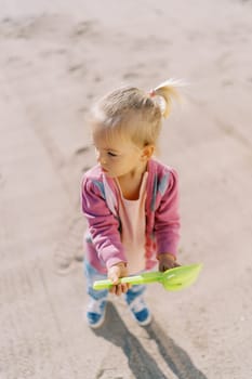 Little girl with a toy shovel in her hands stands on the sand and looks away. Top view. High quality photo