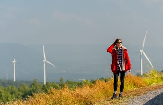 Caucasian woman with red coad and sunglasses walk along the roadside near the meadow and wind turbines or windmill also look to ride side.