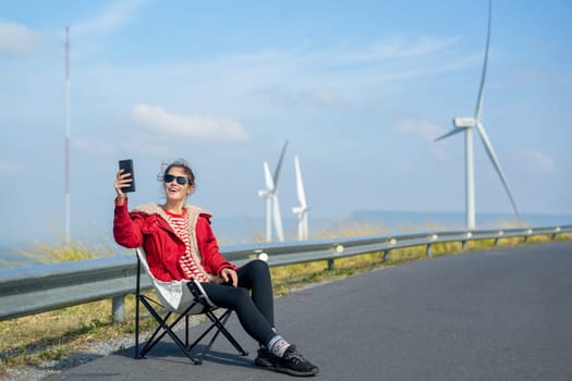 Caucasian woman with red coat and sunglasses sit on chair on roadside near wind turbines or windmill also use mobile phone to selfie with beautiful view on the mountain.