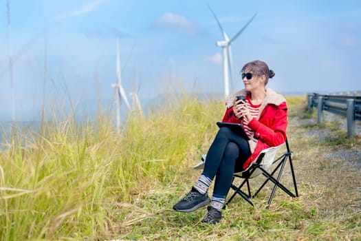 Caucasian woman sit on chair near meadow with blue sky and wind turbines or windmill on the back and she also hold a cup of coffee with happiness.
