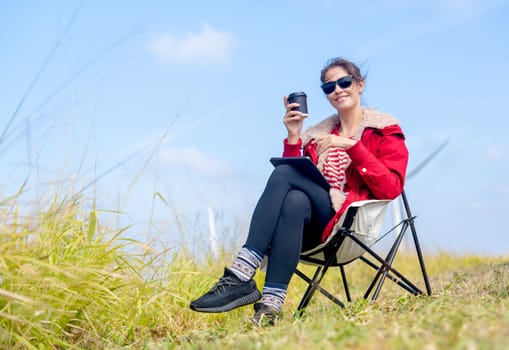 Caucasian woman sit on chair near meadow with blue sky and wind turbines or windmill on the back and she also hold a cup of coffee and look at camera.