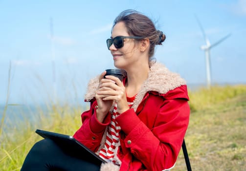 Close up Caucasian woman hold a cup of coffee and look to left side and sit near meadow and wind turbines on the background.