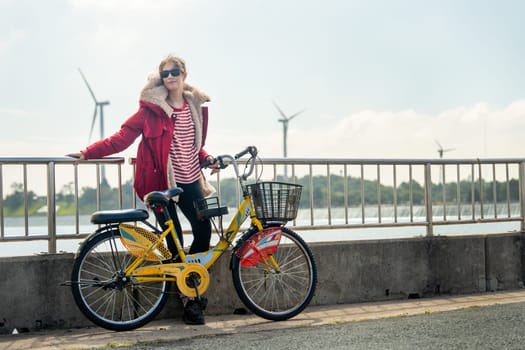 Caucasian woman lean to fence also hold hand of bicycle stand in front of wind turbines or windmill near water reservoir with warm morning light.
