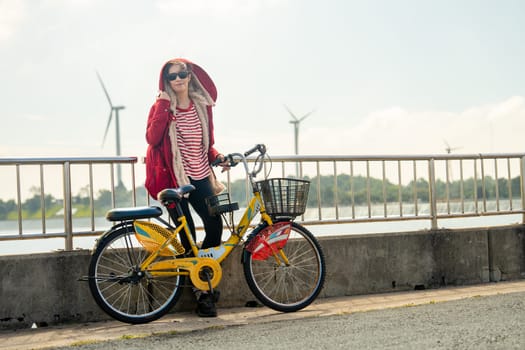 Woman lean with red coat to fence also hold hand of bicycle stand in front of wind turbines or windmill near water reservoir with warm morning light.