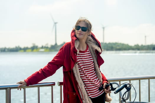 Close up Woman with red coat and sun glasses lean to fence also hold hand of bicycle stand in front of wind turbines or windmill near water reservoir