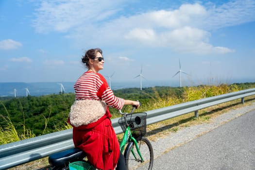 Back of woman with sunglasses stand after ride the bicycle on road on the mountain near row of wind turbines or windmill and she look happiness.