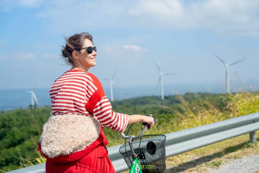 Close up woman with sunglasses stand after ride the bicycle on road on the mountain near row of wind turbines or windmill and she look happiness.