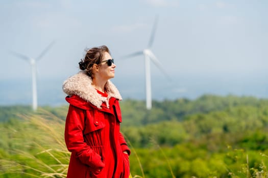 Caucasian woman with red coat and sunglasses stand with smiling and look to right side in front of wind turbines or windmill on the mountain.