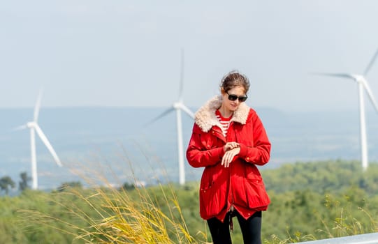 Caucasian woman with red coat look at her watch and stay on mountain with windmill or wind turbine as background.