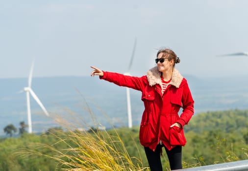 Caucasian woman with red coat stand with smiling and point to left side in front of wind turbines or windmill on the mountain.