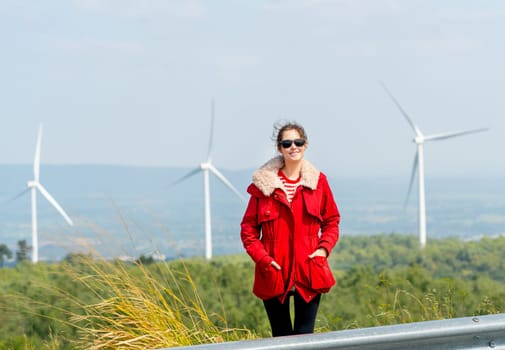 Front view of Caucasian woman with red coat stand with look at camera on mountain near windmill or wind turbine in the back.