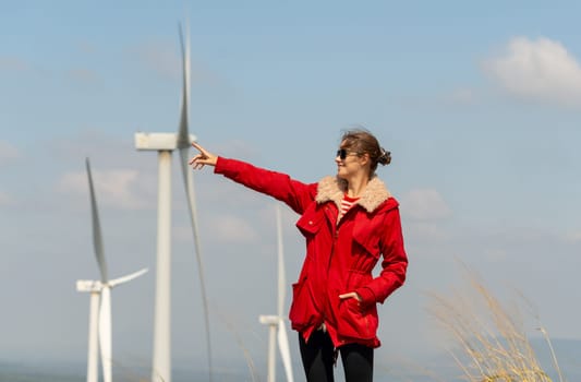 Caucasian woman with sun glasses point to left direction and same position with wind turbine or windmill with blue sky in concept of green energy for good environment.