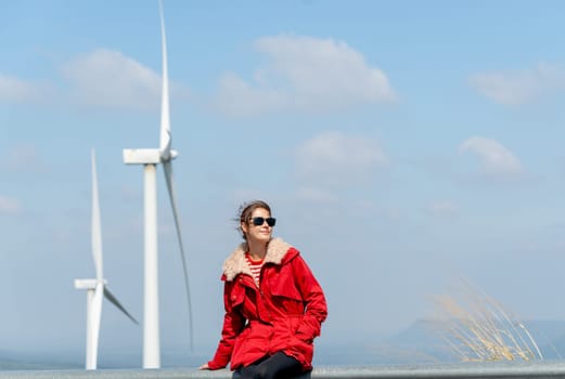 Front view of Caucasian woman with red coat sit on barrier of roadside and use mobile phone on mountain near windmill or wind turbine also look to right side.