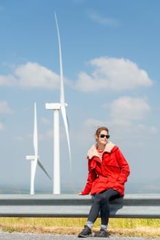 Vertical image of Caucasian woman with red coat sit on barrier of roadside and use mobile phone on mountain near windmill or wind turbine also look to right side.