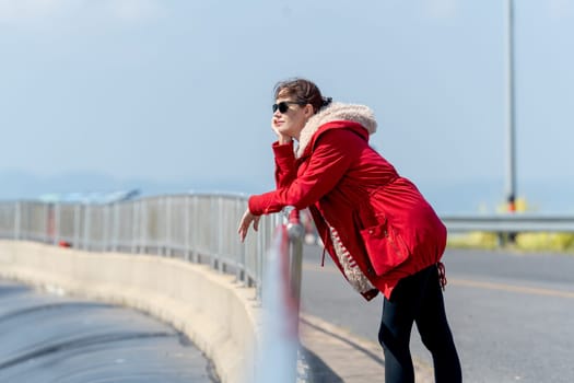 Side view of Caucasian woman with red coat and sun glasses lean to fence and look forward with day light and look cool.