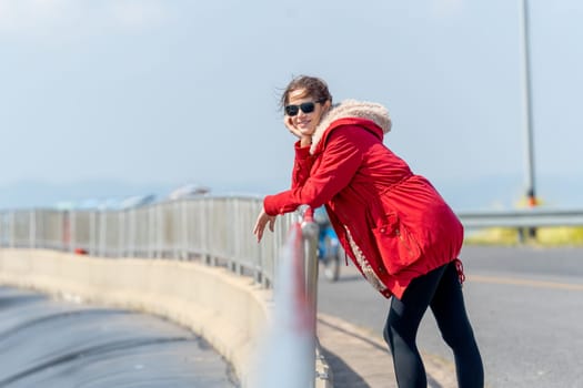 Woman with red coat and sun glasses lean to fence and look at camera with smiling with day light and look cool.
