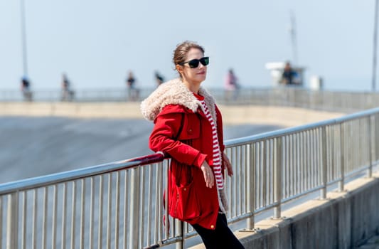Caucasian woman with red coat and sun glasses lean to fence and look to the right with day light and look cool.