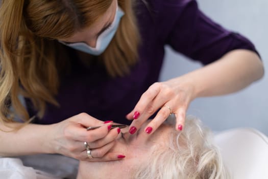 A blonde woman with short hair is lying on a bed in a beauty salon. She is covered with a white quilt. The beautician removes hair from around the client's eyes