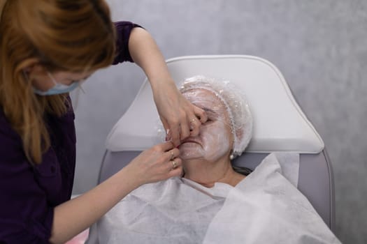 A beauty salon worker carefully wipes a client's face with a cotton swab. The beautician is wearing a purple uniform. The client of the salon is an elderly woman.