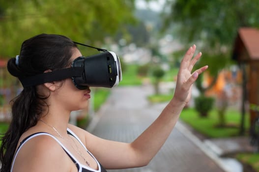 Close-up of woman in blouse and suspenders with virtual reality goggles interacting in a video game next to a wooden house. High quality photo